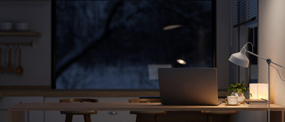 A laptop on a dining table in a contemporary kitchen at night with a dim light from a floor lamp.
