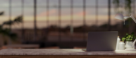 A laptop computer, a table lamp, and a potted plant are on a table in a modern dark office at sunset