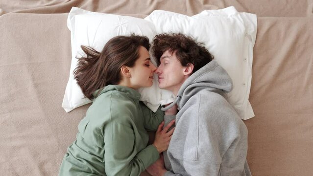 A romantic couple lies in bed, embracing each other. The woman wears a green top, the man a grey hoodie. The cozy bedroom setting adds a relaxed atmosphere. Slow Motion. 