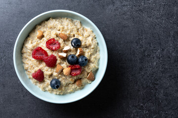 Oatmeal porridge with raspberries, blueberries and almonds in bowl on black background. Top view....
