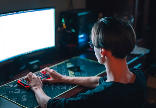 Young programmer at a computer in a dark room. A young man typing on a keyboard, viewed from the back. A teenager sits at a computer at night.