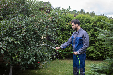 Gardener in uniform holding hose and watering trees and grass on the backyard	
