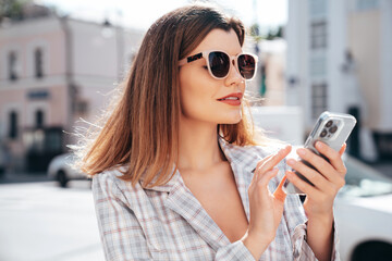 Young beautiful woman in nice trendy white jacket suit. Sexy smiling model posing in street. Fashionable female outdoors. Cheerful and happy. Holds smartphone, uses phone apps, looks at screen