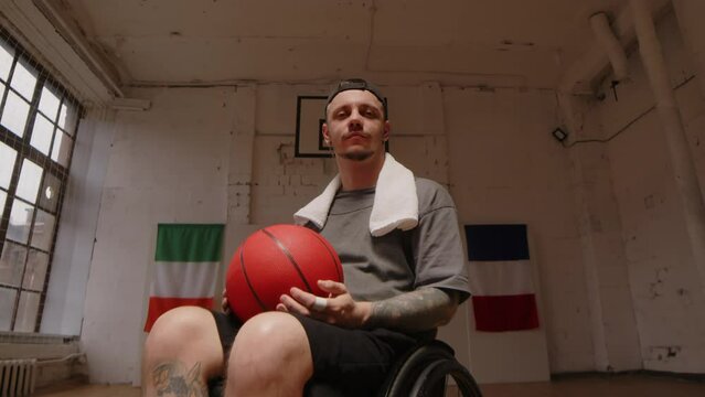 Dolly-in portrait of tattooed young Caucasian man in wheelchair posing for camera on empty indoor basketball court with white brick walls and parquet floor