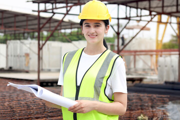 Portrait of young happy beautiful women engineer worker with safety vest and helmet holds blueprint at construction site. Smiling female architect working at ground level of building site workplace.