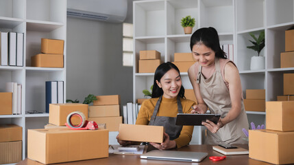 Two Asian women packing boxes and using a tablet in a home office. Concept of small business and e-commerce.