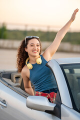 Exuberant young woman with curly hair waves joyfully from a convertible, wearing headphones and...