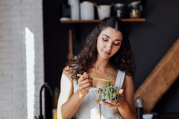 Attractive young woman is eating salad of sprouted greens in kitchen. Curly-haired brunette girl in linen sundress holds fork and transparent bowl with micro greenery.