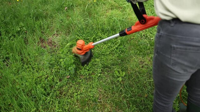 Female gardener using electric string trimmer to trim grass in garden. Lawn care in backyard