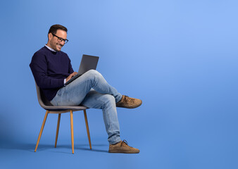 Confident young businessman in eyeglasses using laptop while sitting on chair over blue background