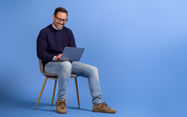 Happy male freelancer working on project over laptop while sitting on chair against blue background