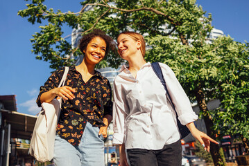 Two cute female students in casual clothes with backpacks walk along the city street and...