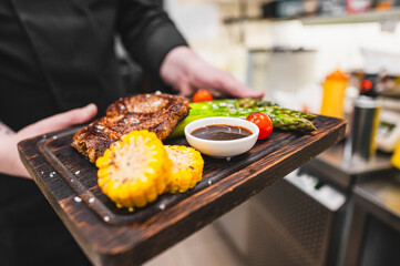 Close-up of hands holding a wooden tray with grilled steak, asparagus, corn on the cob, and a small bowl of sauce. - Powered by Adobe
