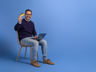Portrait of cheerful young man with laptop and credit card sitting on chair against blue background