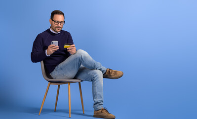 Businessman shopping online with cellphone and credit card while sitting on chair on blue background