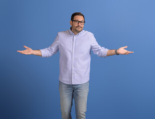 Confused businessman spreading hands and shrugging shoulders while standing against blue background