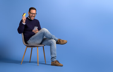 Excited young man with credit card and mobile phone screaming happily on chair over blue background