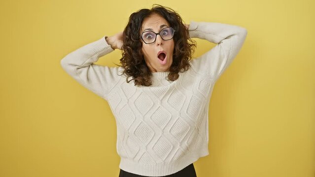 Shocked! crazy-scared middle-aged hispanic woman with glasses, standing hands-on-head, mouth open wide in surprise against a yellow cut-out background
