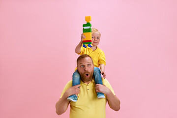 Cheerful father gives his son shoulder ride while he proudly holds up her colorful building block...