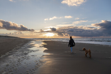 Female with her pet dog walking along the shoreline of a beach at sunset
