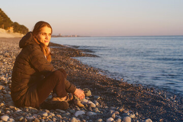 Young smiling woman relaxing in the evening on a pebble beach by the sea and looking into the distance at sunset in spring
