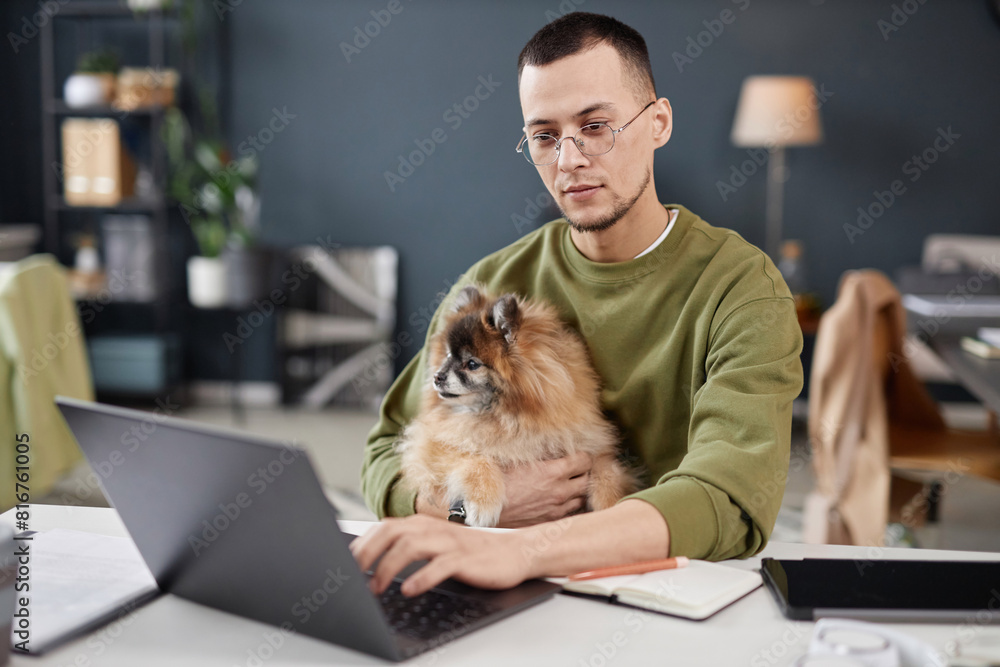 Poster portrait of young man using computer and holding cute dog in lap while working in pet friendly offic