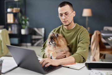 Portrait of young man using computer and holding cute dog in lap while working in pet friendly...