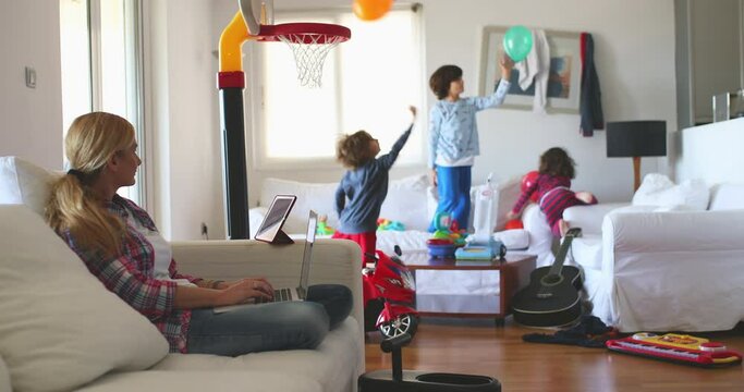A focused mother works on her laptop on the sofa, while her playful children engage in active games around her, depicting the challenges of remote work with kids at home.