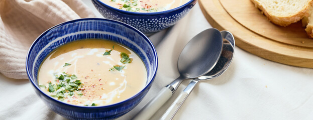 Two bowls of creamy potato soup, spoons, napkin and wooden cutting board with a loaf of bread and cut pieces.