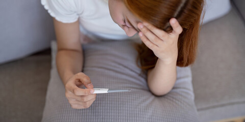 Naklejka premium Young Asian woman with fever checking her temperature with a thermometer at home. Concept of illness and healthcare