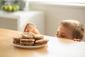 A boy is peeping at an appetizing stack of freshly baked chocolate cookies on the table.