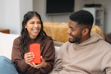 Smiling Indian woman enjoying gift given by American African  man sitting on sofa in living  room at home