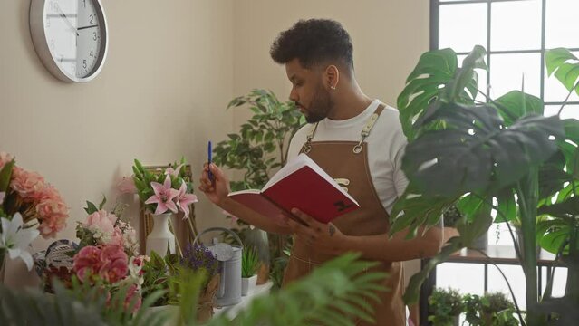 A man in a flower shop arranges bouquets while consulting a red notebook, surrounded by plants and decor.