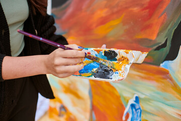 Female painter hand dips paintbrush into palette of colorful paints for live painting of picture...