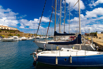 Vista detallada del puerto de Stintino en Cerdeña, Italia, durante la primavera. Varios veleros y yates están atracados en el puerto, rodeados de muelles de piedra y edificios mediterráneos.  © jocaja