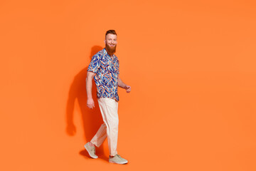 Full length photo of positive optimistic guy wear print shirt white trousers go to empty space isolated on orange color background