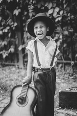 a little guitarist in a hat stands on the street leaning on his guitar and laughs
