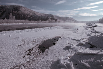 A foggy winter river with mountains, trees on shore, and clouds in sky