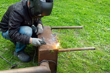 A man welder in a welding mask and gloves welds metal using an arc welding machine outdoors, sparks...