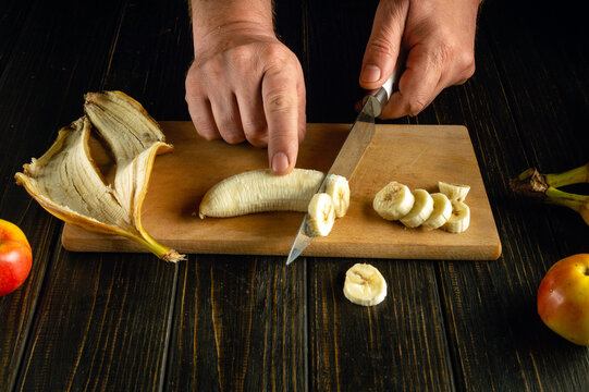 Slicing a ripe banana with a knife in the hands of a man for preparing a fruit menu on the kitchen table. - Powered by Adobe