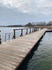 Wooden pier extending into a calm lake under a cloudy sky, surrounded by bare trees and reeds,...