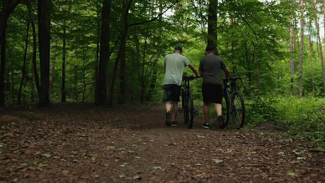 Two friends slim guy and unsportsmanlike overweight man walk roll hardtail mountain bikes nearby in forest path, view from back. Adult men carry bikes nearby. Cyclists took break, weight loss training
