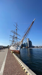 A majestic sailing ship docked at a modern harbor. Tall masts and rigging against a clear blue sky,...