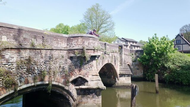 Woman stood on medieval arched Bishops bridge looking over parapet into river Wensum