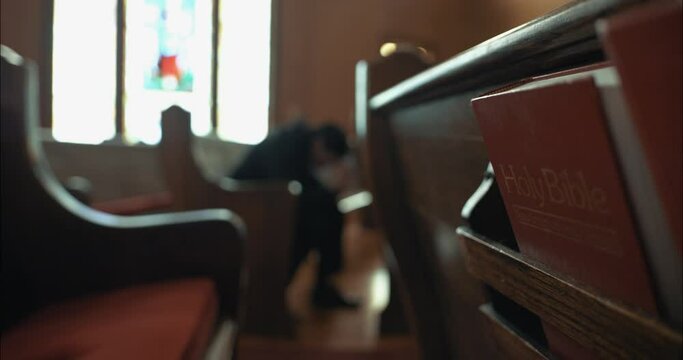 Young man in black suit praying in church in cinematic slow motion with folded hands with bible in pew.