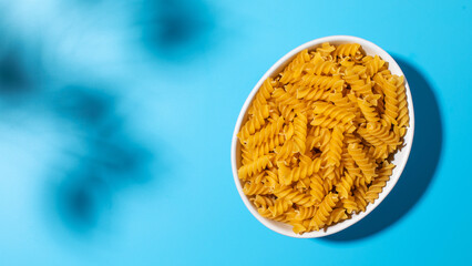 Uncooked fusilli pasta spirals in a white bowl on a blue background with shadows