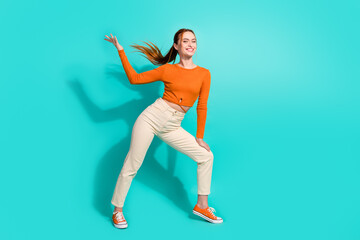Full size photo of adorable girl with fluttering ginger hair dressed orange shirt raising hand up isolated on teal color background