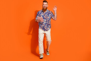 Full length photo of friendly nice guy with red beard wear print shirt pants hold tablet in office isolated on orange color background
