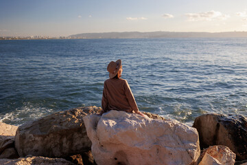 back view. a young girl wearing a coat and hat sits on a high stone and looks at the beautiful view...