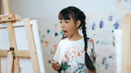Playful student painted or draw canvas at stained wall in art lesson. Asian girl wearing white...